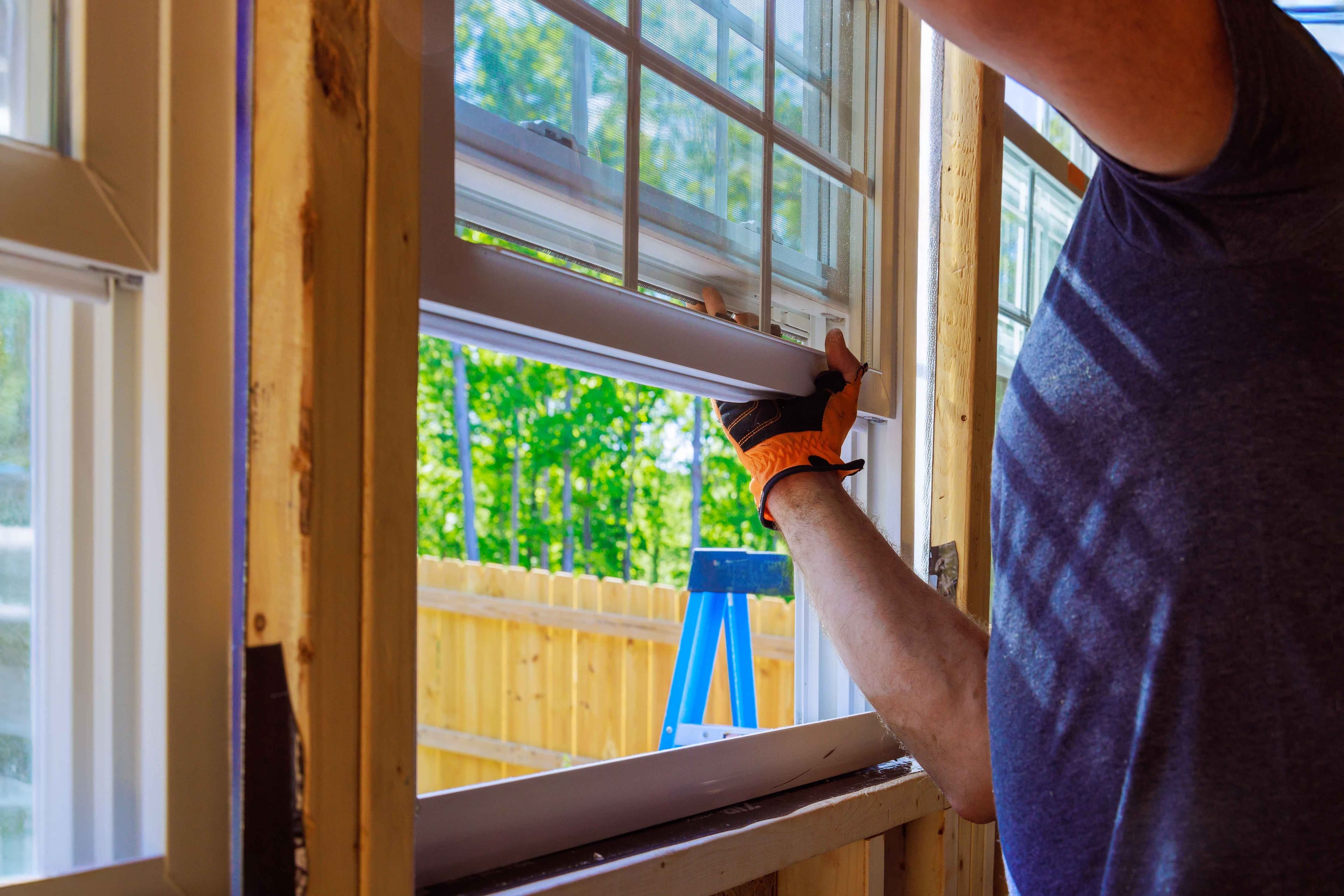 A gloved hand fitting a double-hung window with its screen into a wood-framed exterior opening during window replacement.