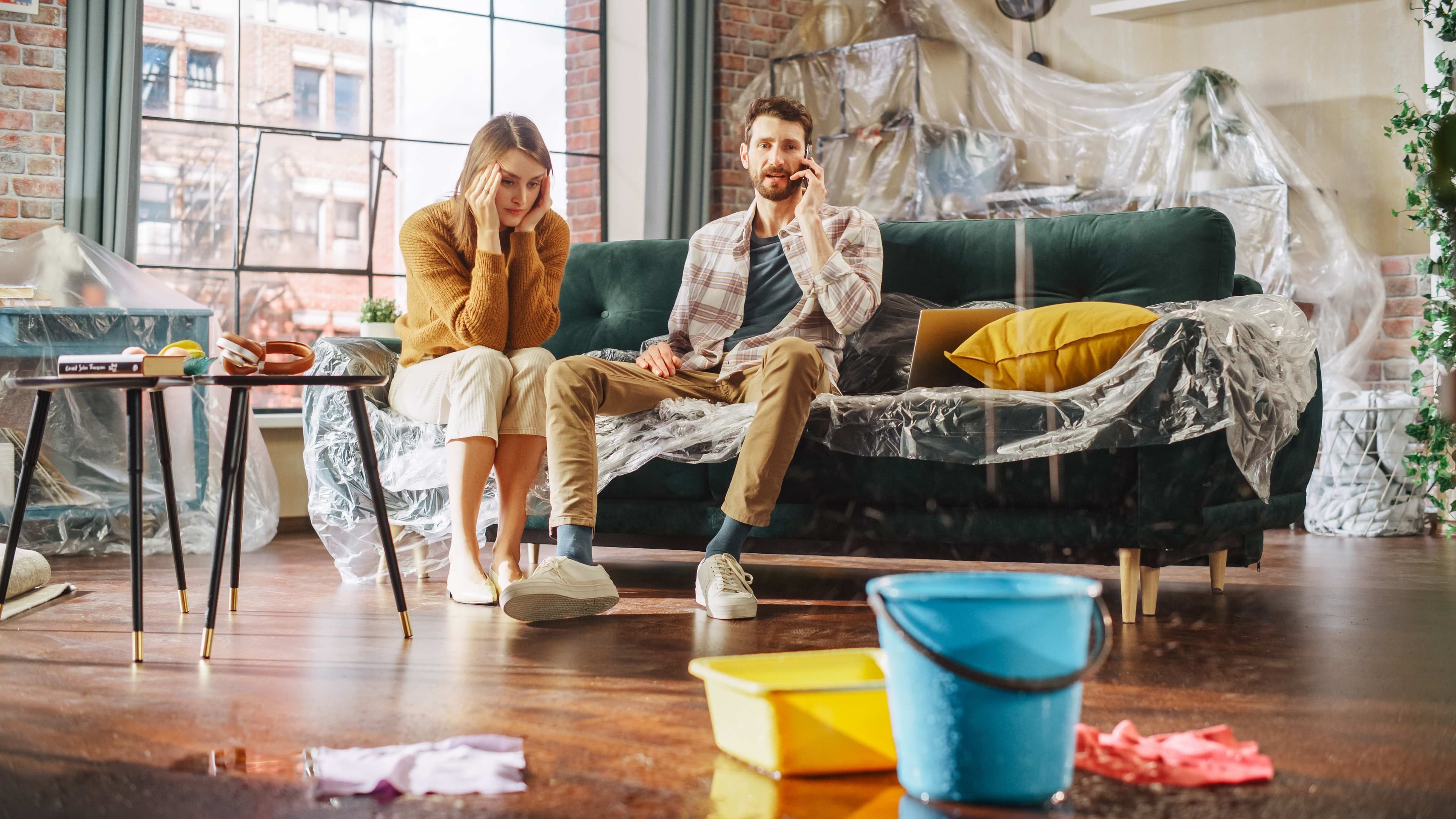 A woman resting her head in her hands and a man on a phone call sitting on a plastic-covered sofa in a room with buckets on the floor