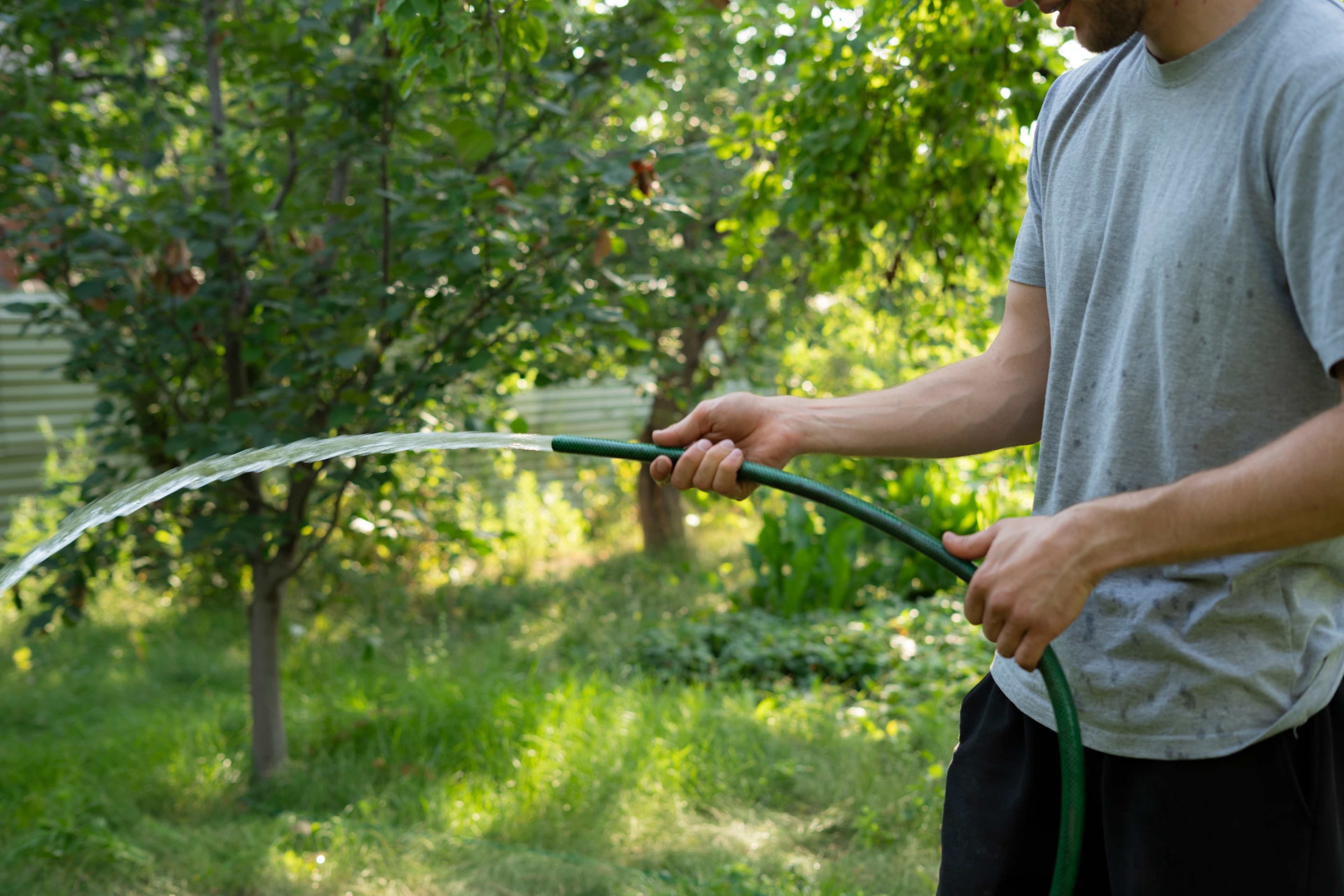 A person holding a green garden hose and watering plants and grass in a backyard