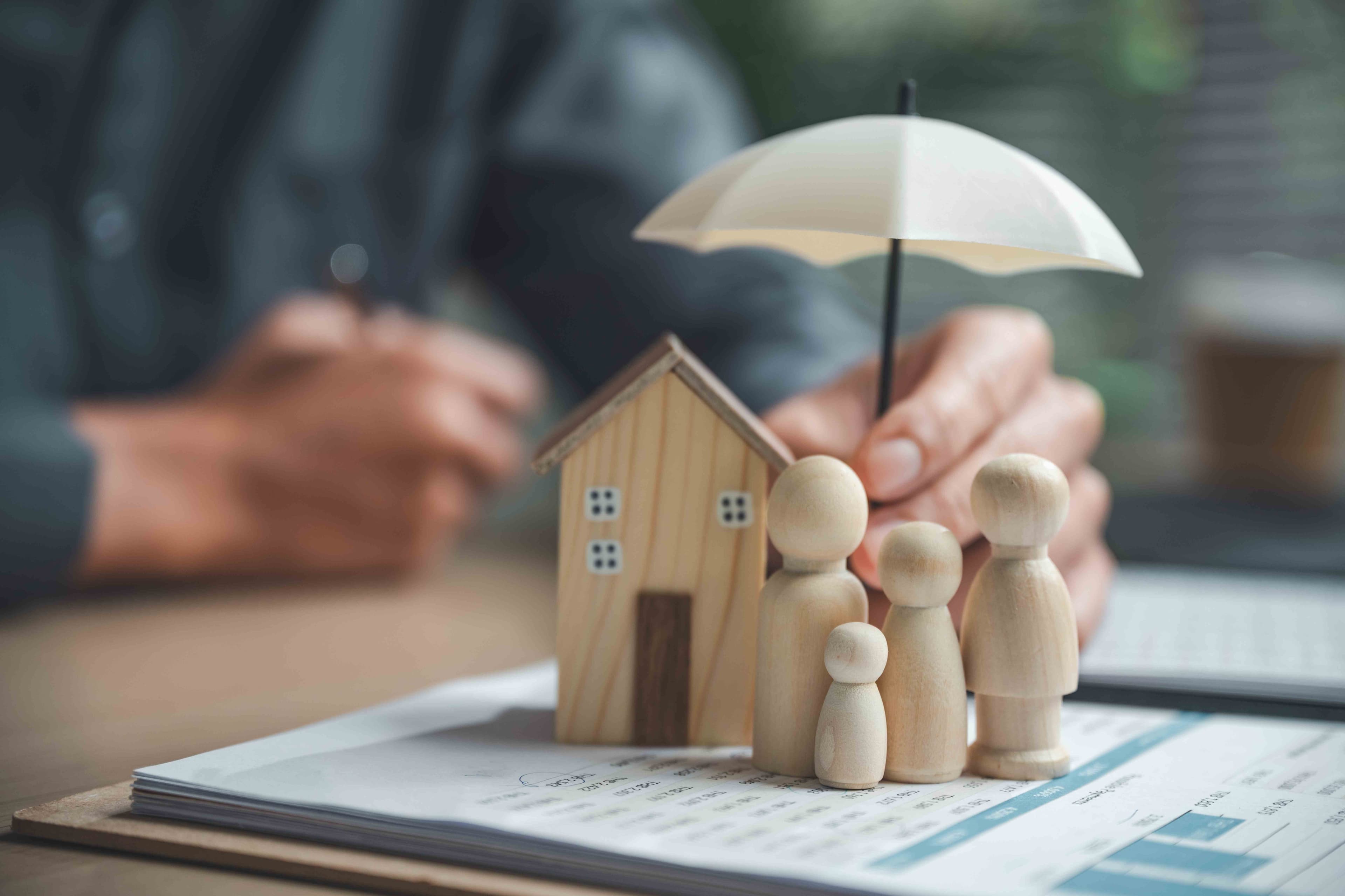 Wooden figures of a family and a small house model beneath a toy umbrella held above insurance paperwork