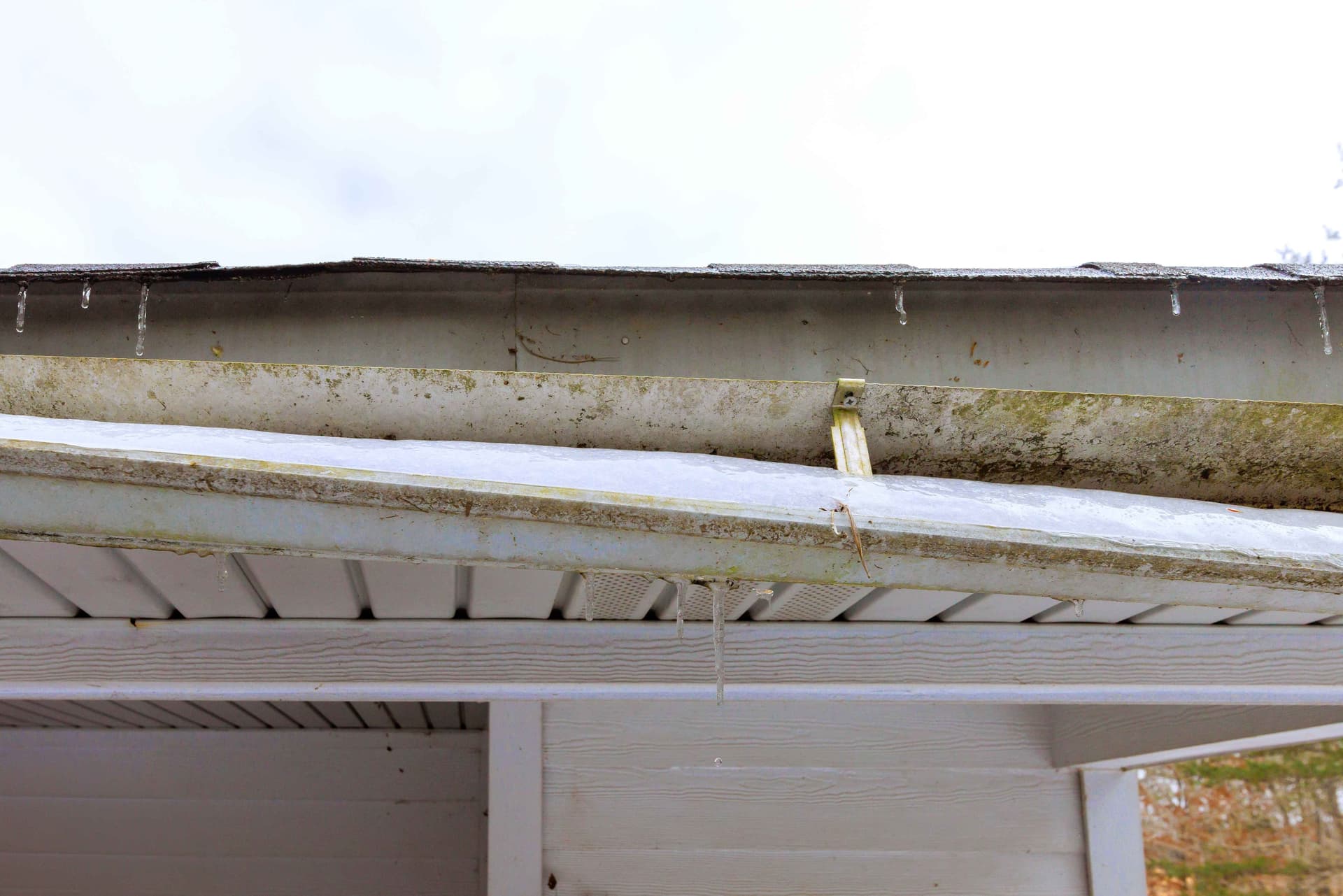 Close-up of dirty gutter with icicles hanging from the edge and debris buildup